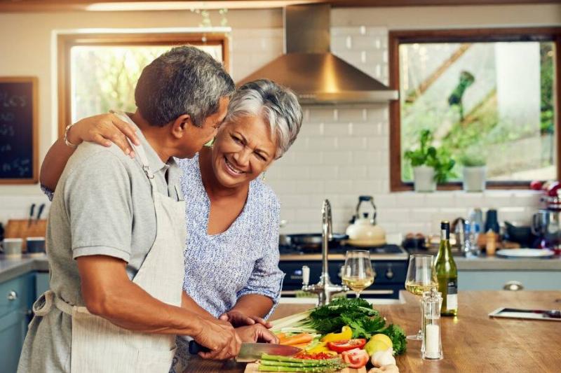 A couple smiling while in the kitchen, the woman putting an arm around the man who's chopping veggies.