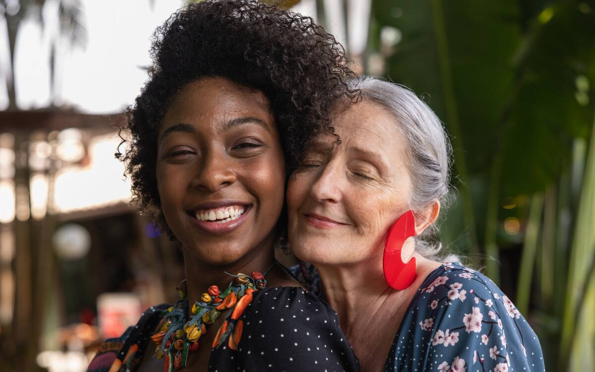 Two women embracing amicably, pressing their heads together, one behind the other, both smiling.