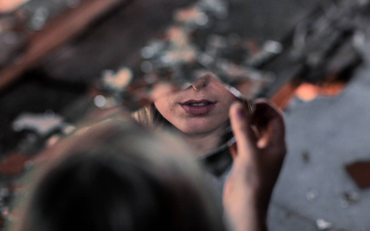 A woman holding up a shattered shard of a mirror, her mouth reflected in it.