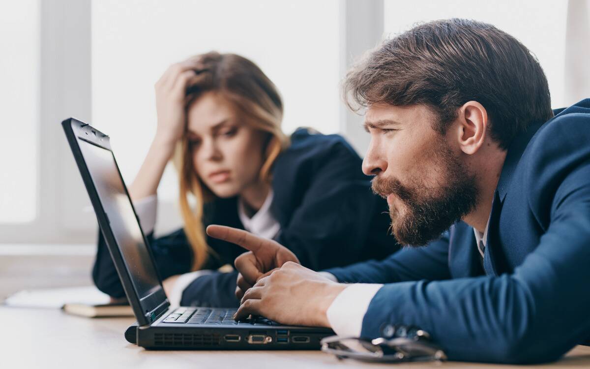 A man pointing at his computer, confused, as a woman slouches tiredly behind him.