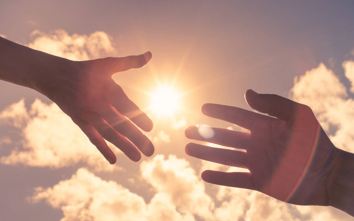 Two hands reaching out for one another in front of the sun.