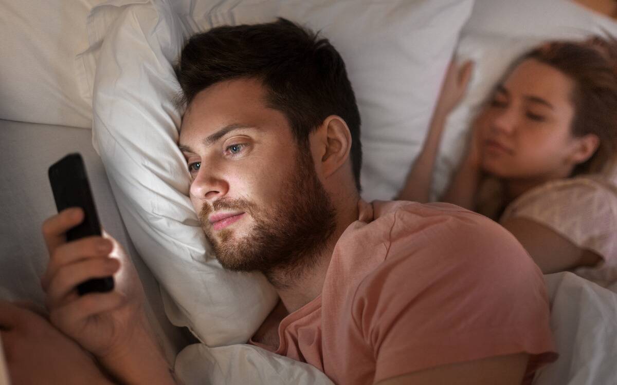 A man looking at his phone while in bed with his girlfriend.