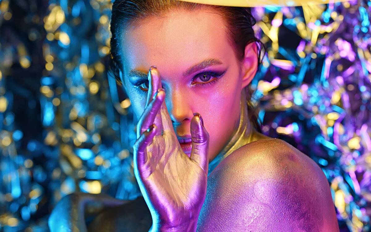 A woman in shiny gold body paint in front of a neon lit, glittery background.