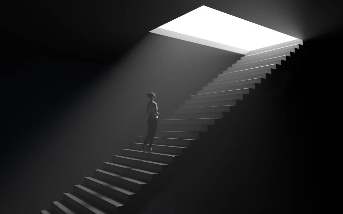 A woman walking up a dark set of stairs toward a square opening of light.