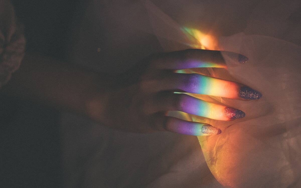 A woman's hand grabbing at some fabric, sporting long, glittery nails, and with a streak of rainbow light shining overtop her fingers.