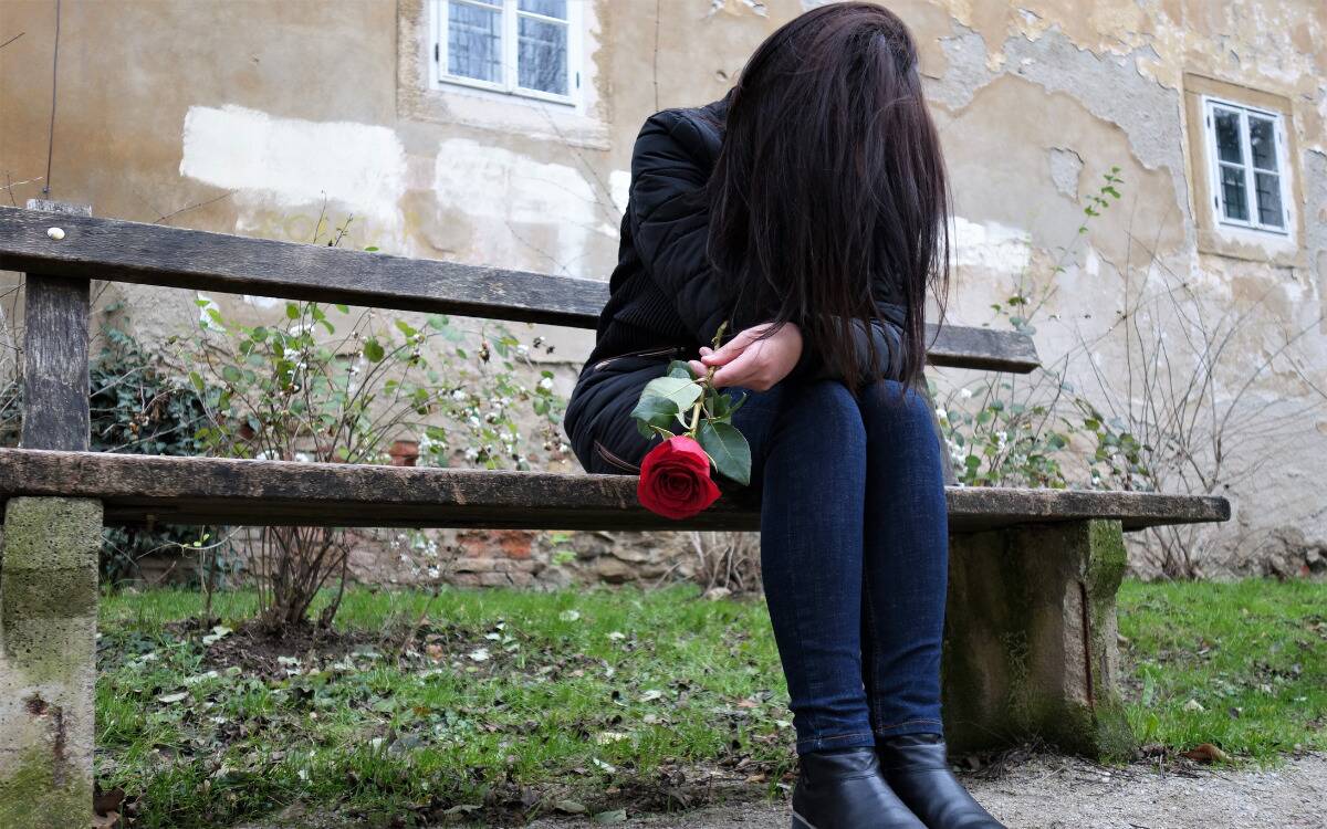 A woman sitting on a bench alone, head hung forward, a rose in her hands.