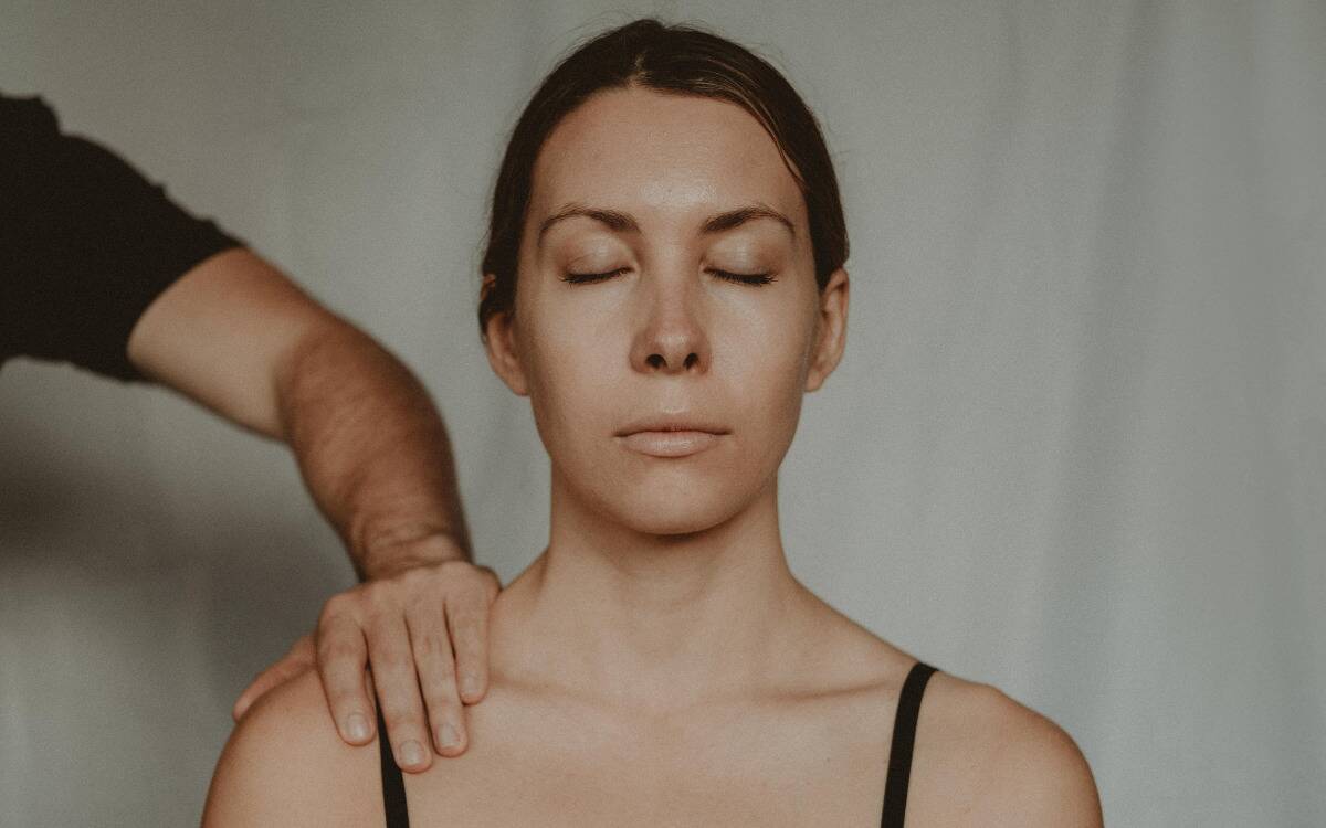 A woman sitting with her eyes closed, an off-screen man placing a hand on her shoulder.