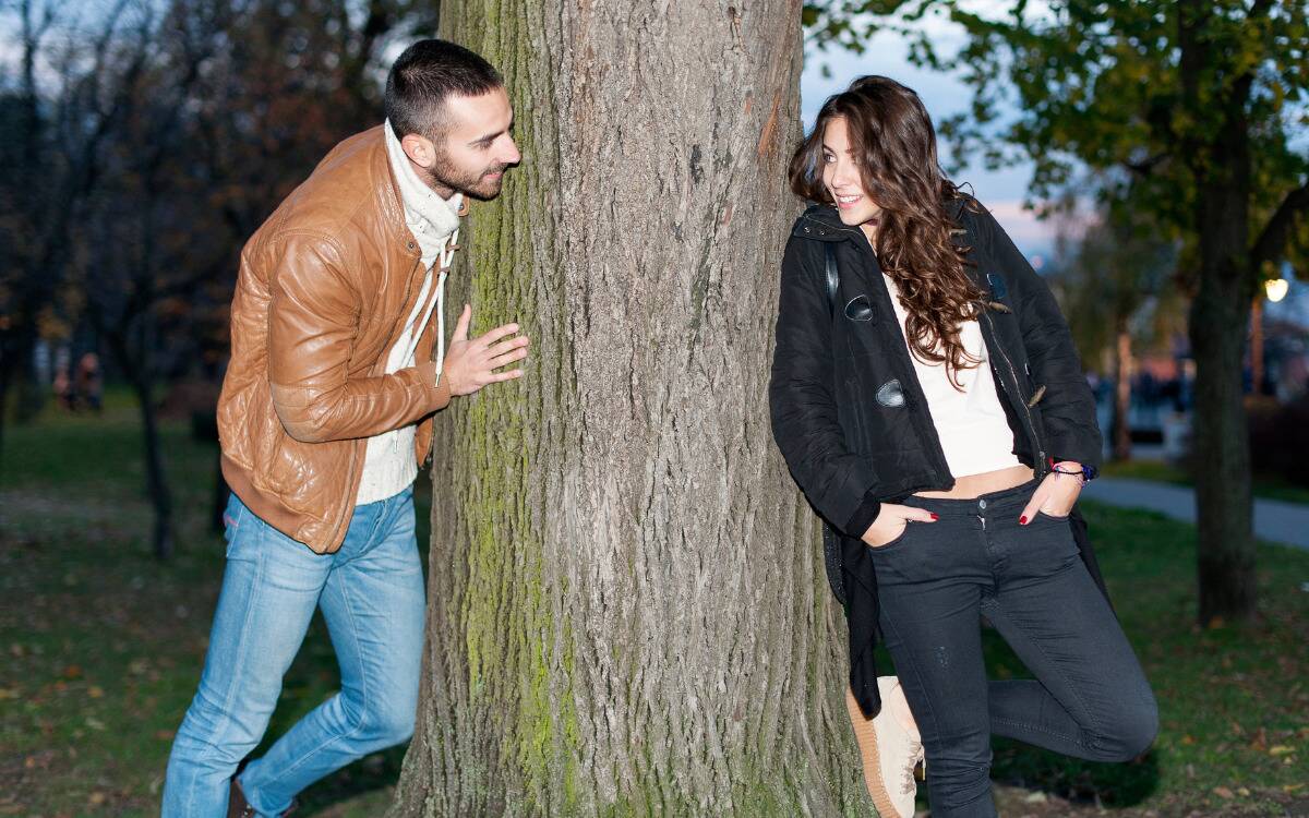 A man and a woman both looking at each other from opposite sides of a tree.