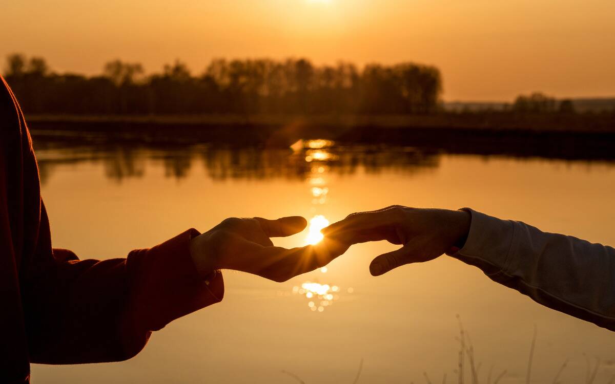 A silhouette of two hands with their fingertips touching in front of a lake at sunset.
