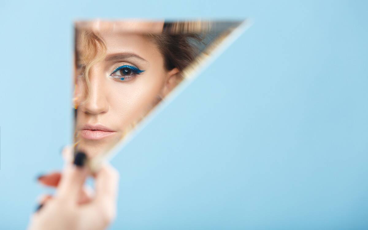 A woman looking in a triangular mirror, wearing bright blue eye makeup.
