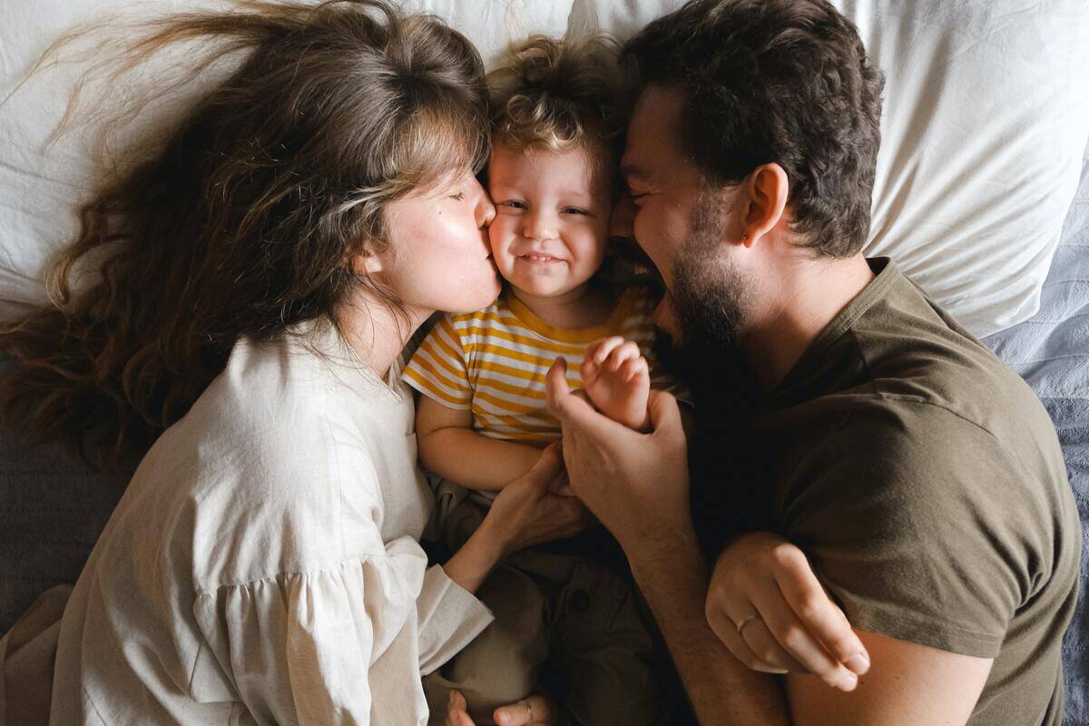 Two parents on either side of their young son in bed, the mom kissing him on the cheek while the dad holds his hand.