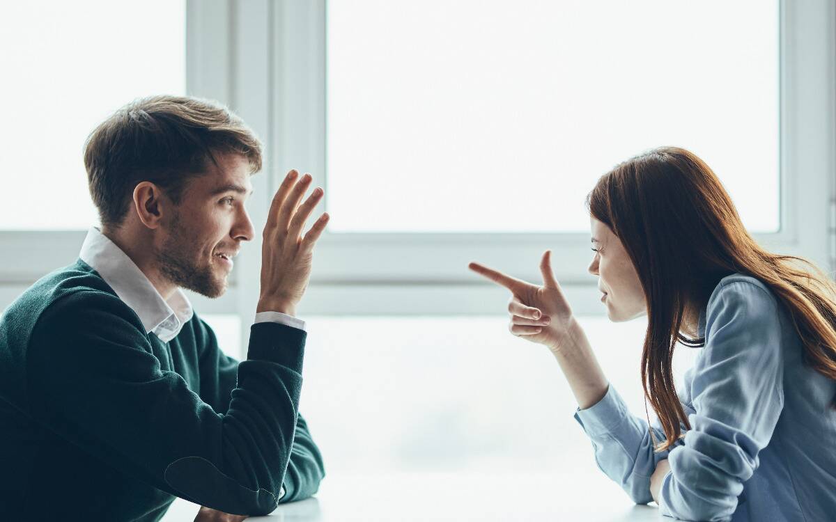 A couple arguing across the table, the woman pointing at the man in an accusatory manner.