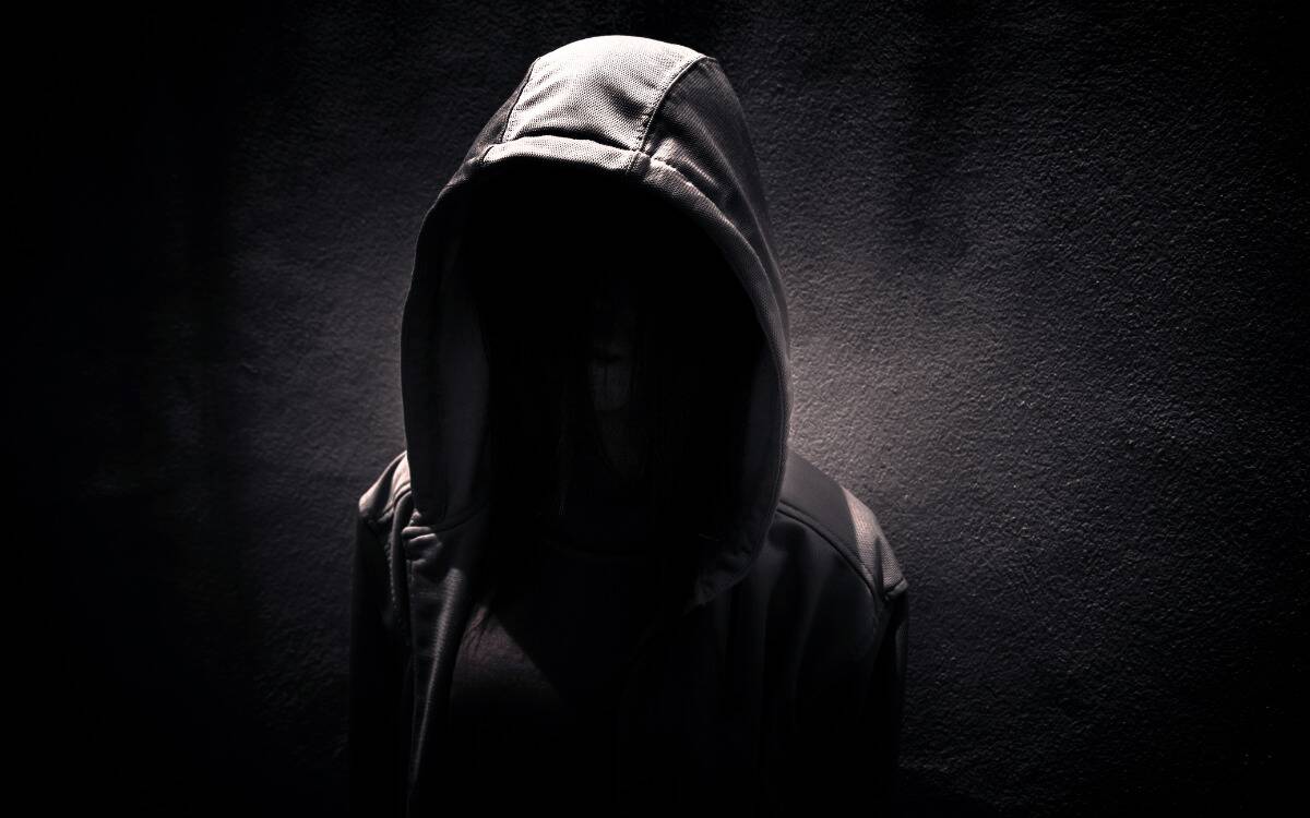 A greyscale  image of a person in a hoodie, their face completely obscured in shadow. 