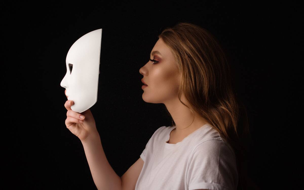 A profile shot of a woman holding up a mask a few inches in front of her face.