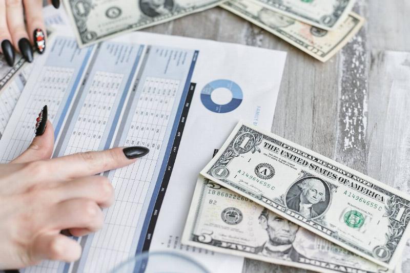 A woman with pointy, black painted nails pointing out banking charts, cash bills on the table around her.