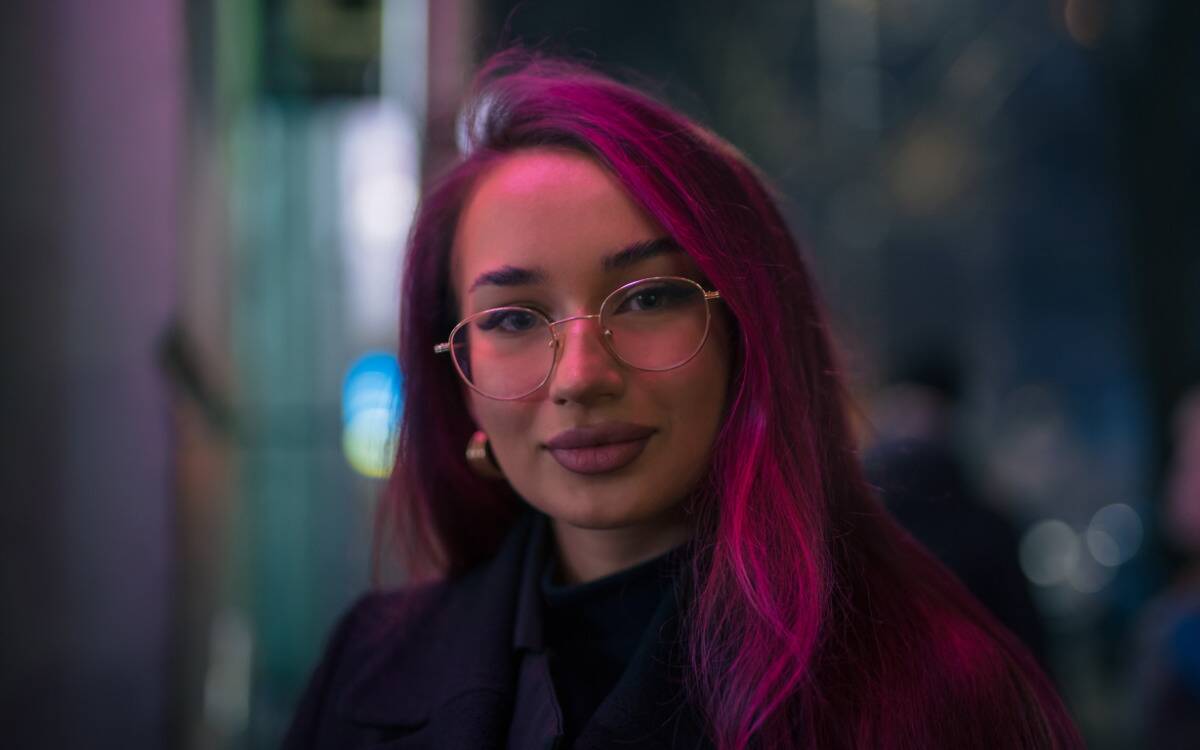 A portrait of a girl with glasses and long pink hair, standing on the street outside.