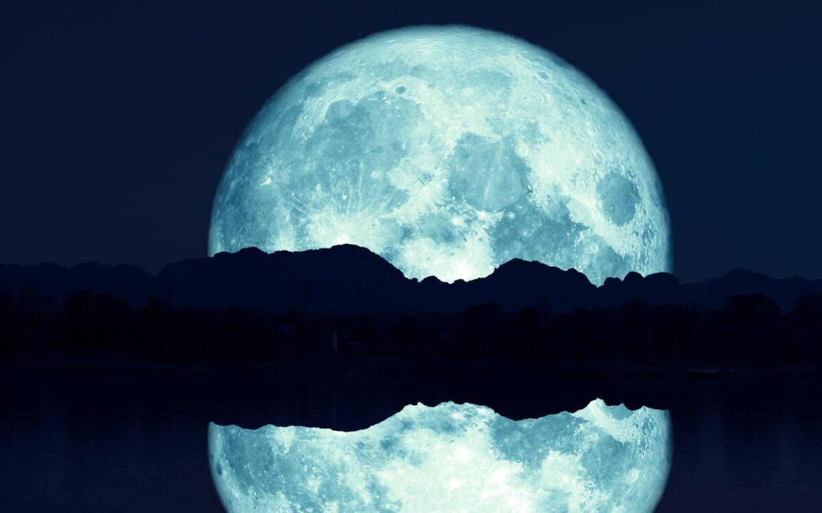 A blue moon rising above a hill range and a body of water, reflected in the water.
