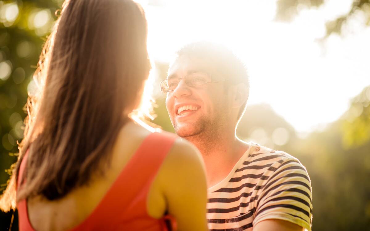 A couple smiling at one another in the sunshine, seen from over the woman's shoulder.