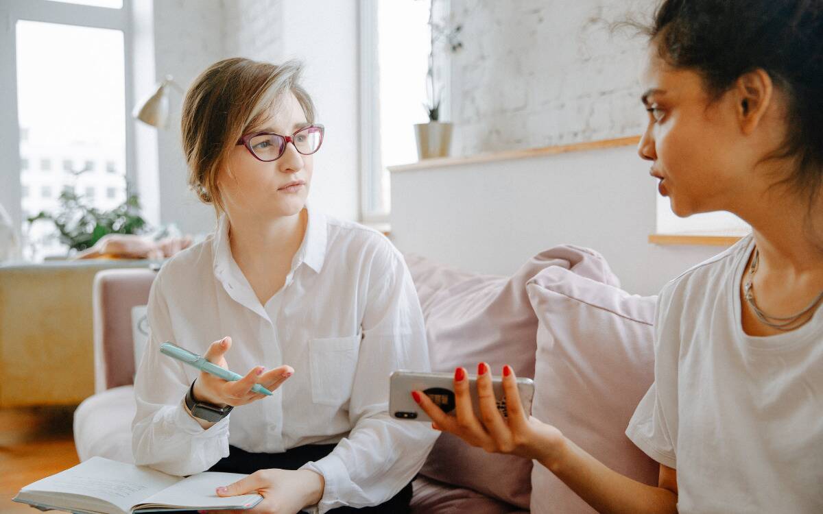 Two women talking while sitting on a couch, one writing things down as they speak.