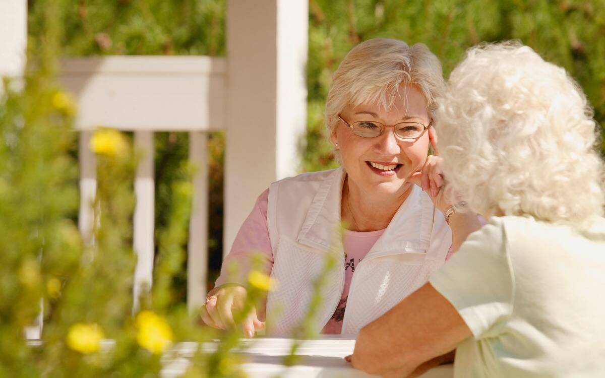 Two older women sitting at a table outside talking with one another, smiling.