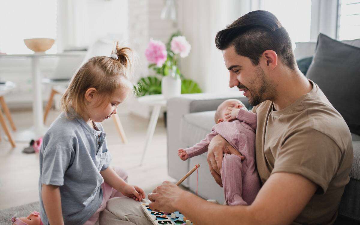 A father playing with a toy with his daughter while also holding a second baby.