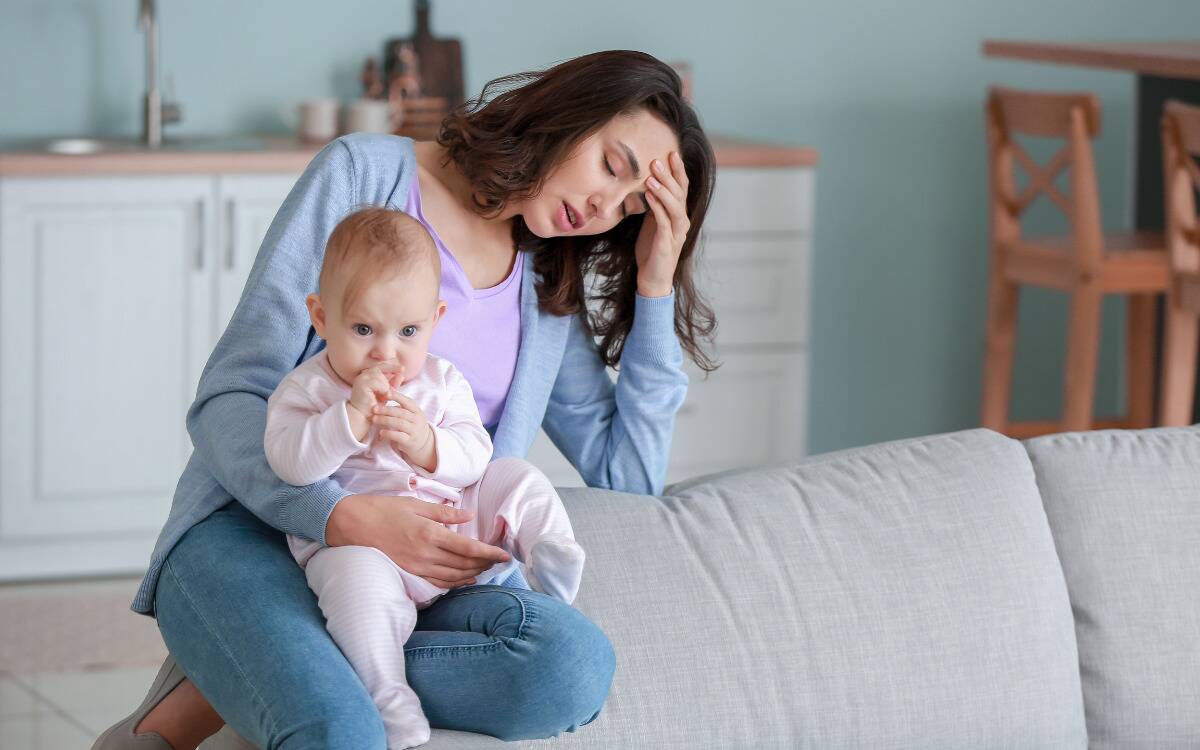 A mom looking tired, leaning on a couch while she's holding her baby.