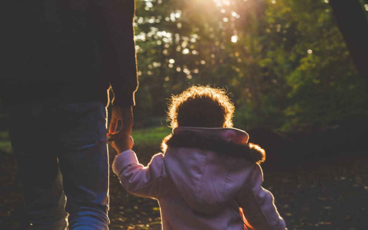 A parent walking with their kid, hand in hand, through a forrest.