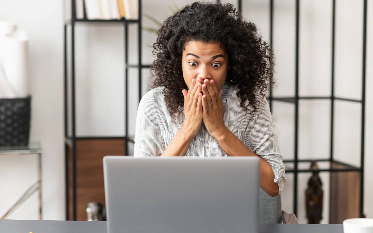 A woman looking at her laptop with a shocked expression, mouth covered by her hands.