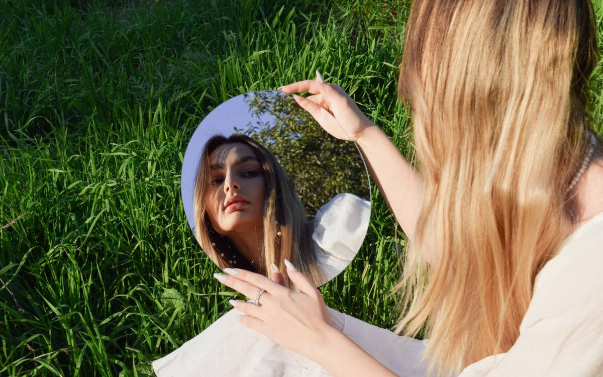 A woman sitting in the grass, looking into a mirror she's holding low to the ground, seeing her reflection.