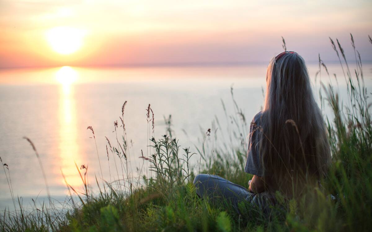 A woman sitting in some tall grass, looking out at the sun set over the water.