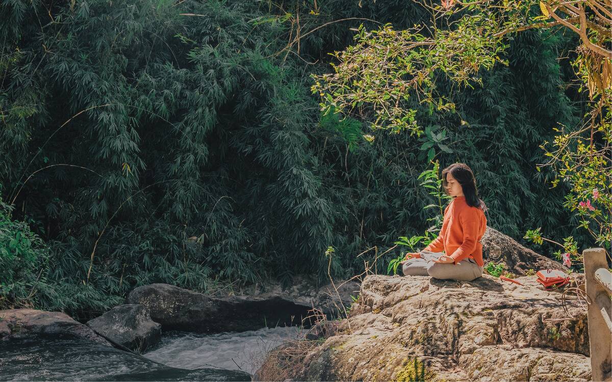 A woman sitting and meditating by a creek.
