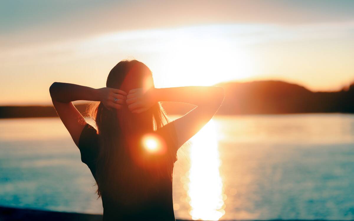 A woman standing with her hands behind her head, facing the sunrise.