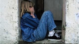 A young girl sitting in a doorway, knees drawn up, covering her face.