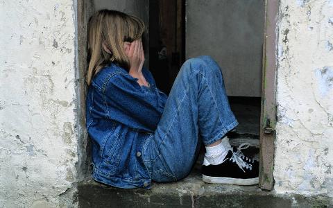 A young girl sitting in a doorway, knees drawn up, covering her face.