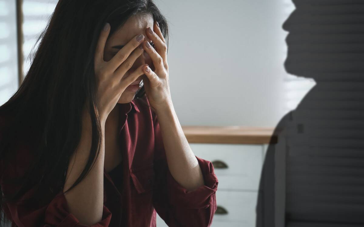 A woman covering her face with her hands as the silhouette of a man looms beside her.