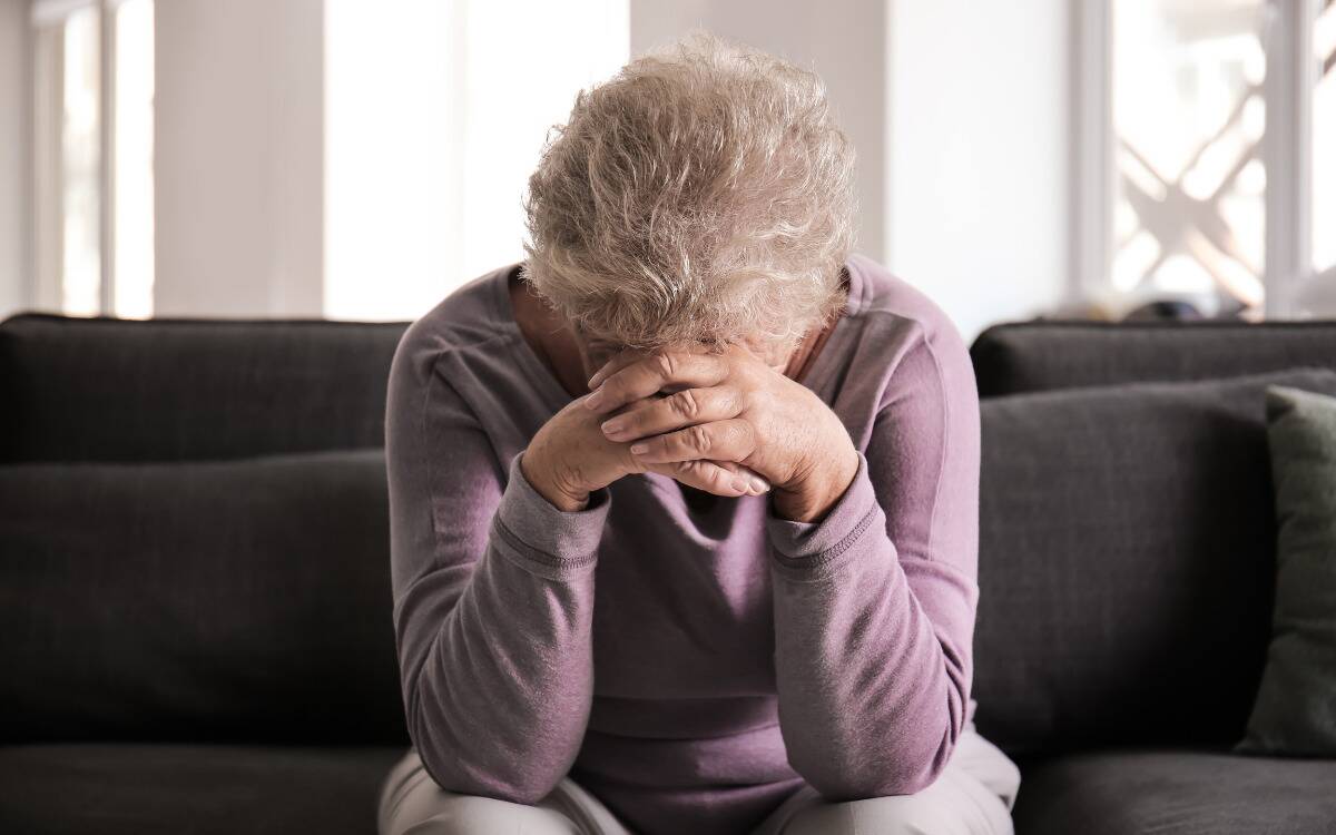 An older woman sitting on a couch, leaning forward, her head in her hands.