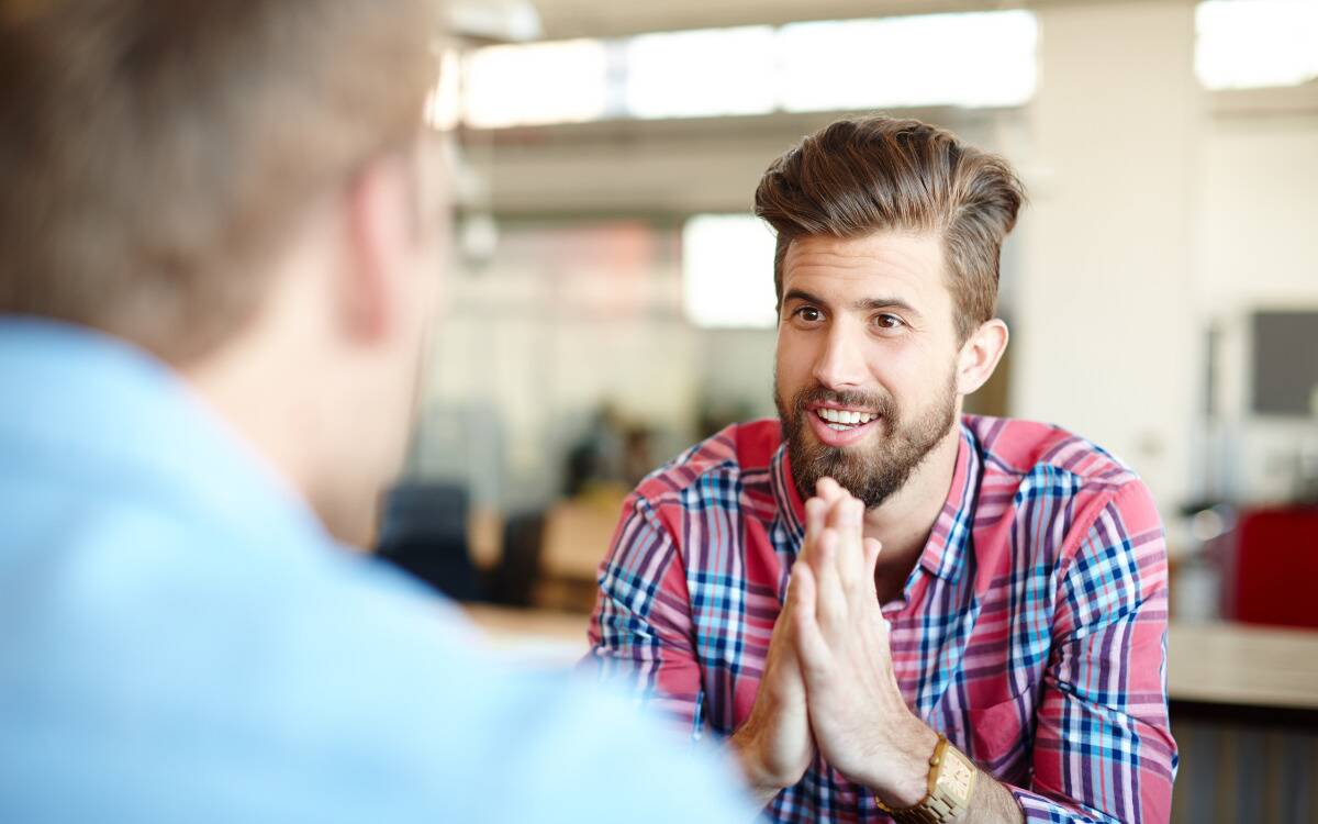 Two men talking, the shot taken over the first man's shoulder. The man he's talking to has his hands pressed together as he listens.