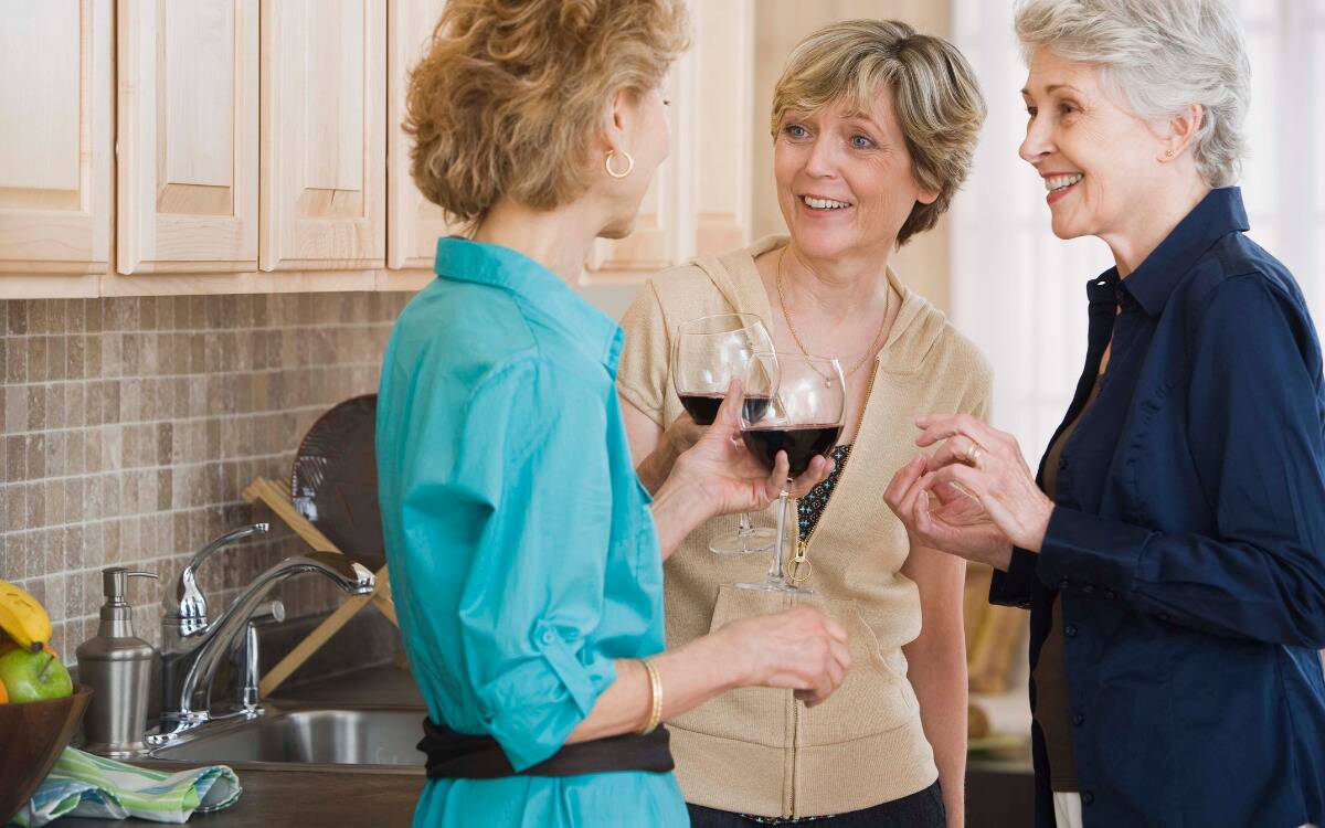Three women talking in a kitchen, holding glasses of wine.