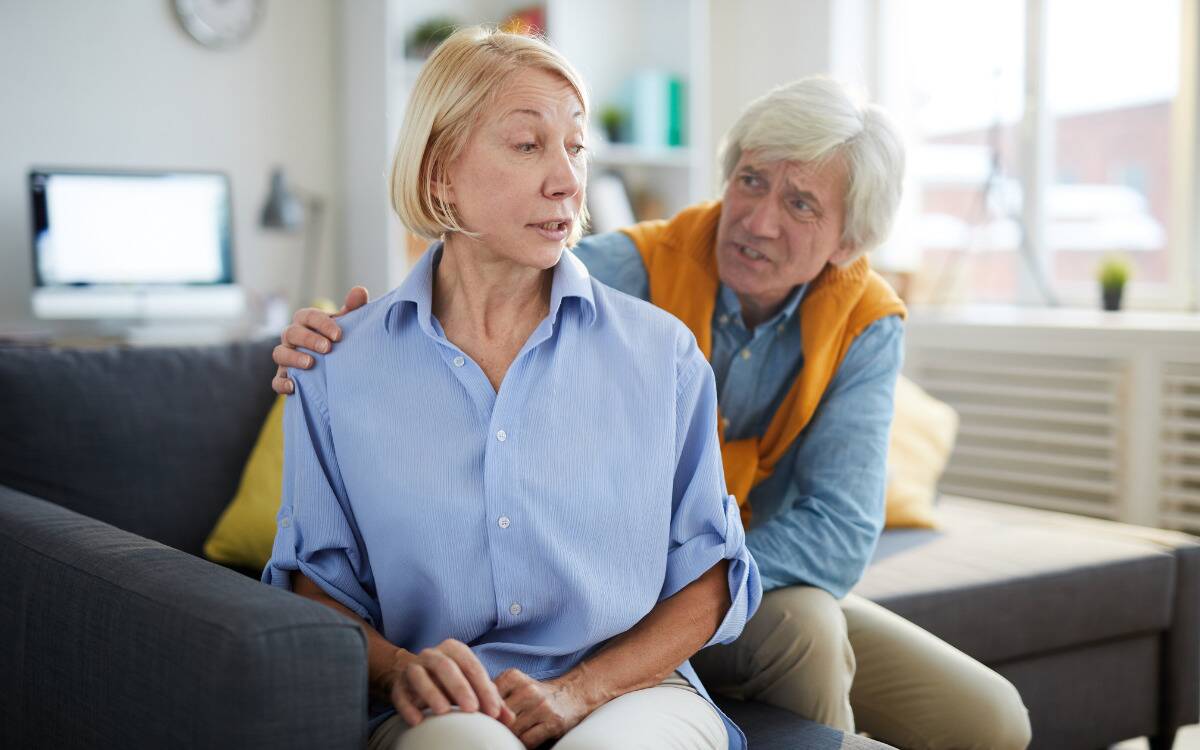 A woman sitting on a couch, facing away from her husband who's trying to apologize to her. She looks unimpressed