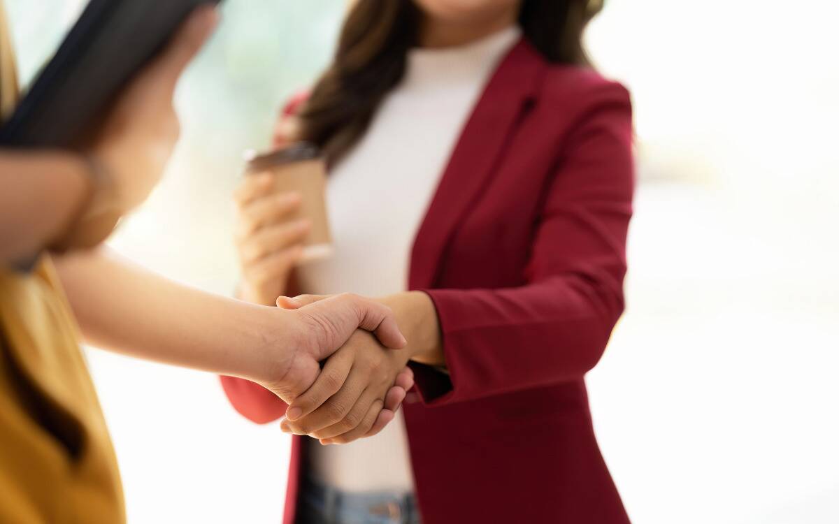 Two women shaking hands, one in a red blazer holding a cup of coffee.