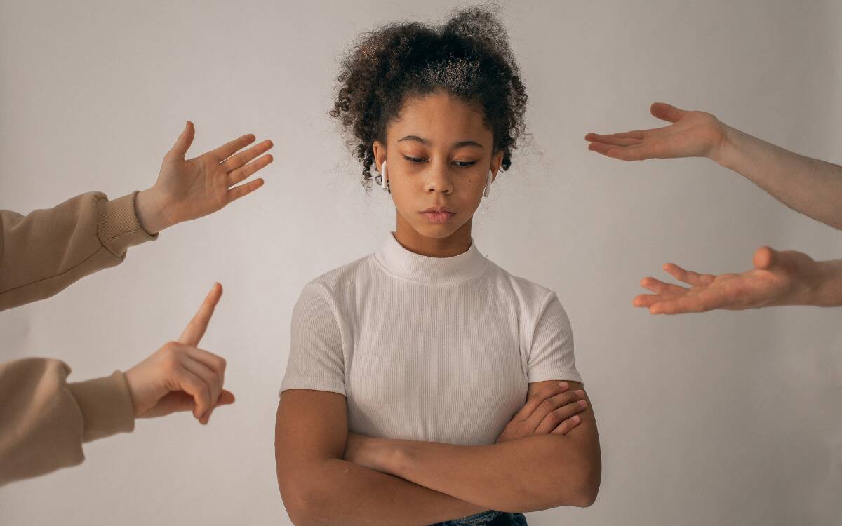 A girl with her arms folded. On either side, there are the hands of people who are talking to her, the hang gestures suggesting yelling or criticism.