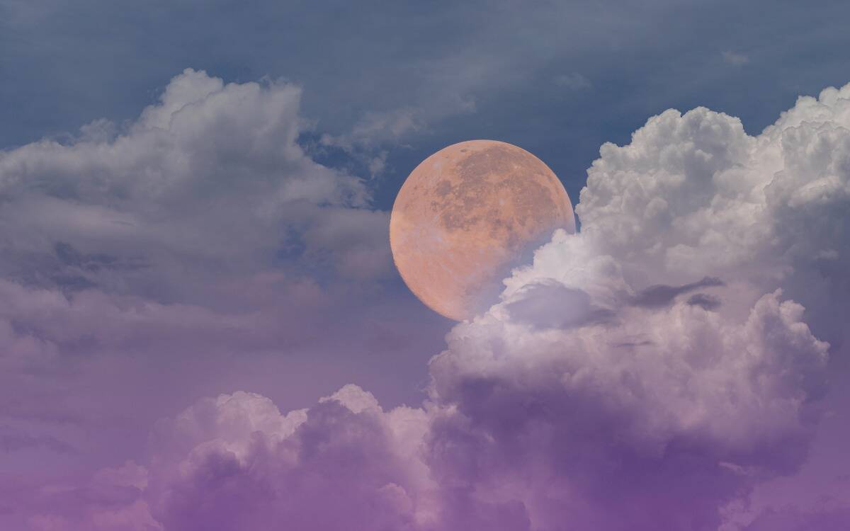 The full moon in the sky, hanging among clouds in a blue/purple sky.