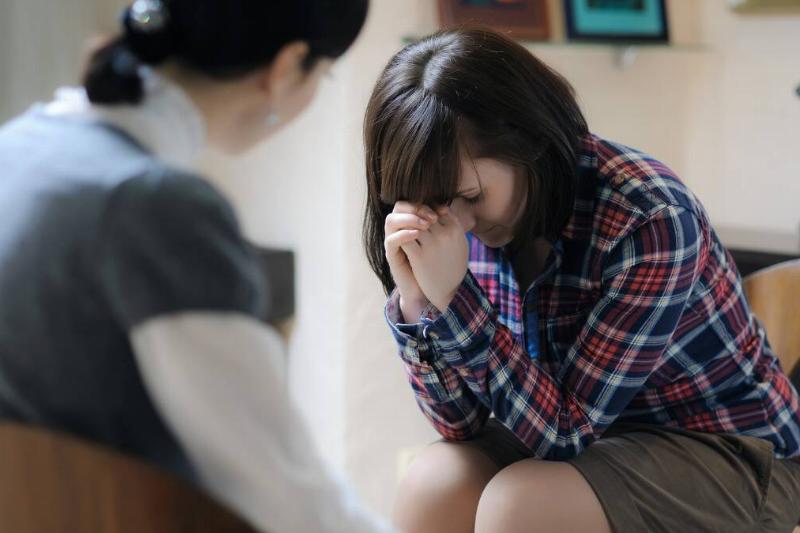 A girl leaned over, forehead in her hands, looking distressed as she speaks to a therapist.