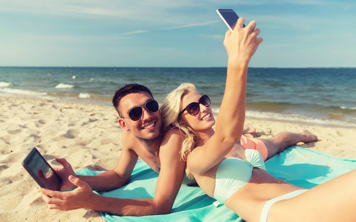 A couple taking a selfie on the beach, the man laying on his stomach while the woman lays back against his shoulder.