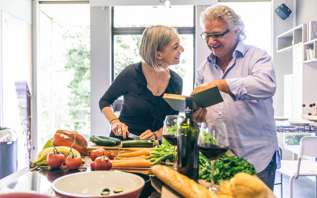 A couple both looking at a cookbook in their kitchen, their counter covered in vegetables.