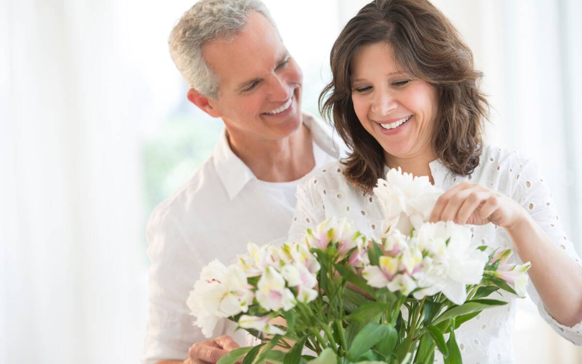 A man looking over a woman's shoulder as she arranges a flower bouquet, both smiling.