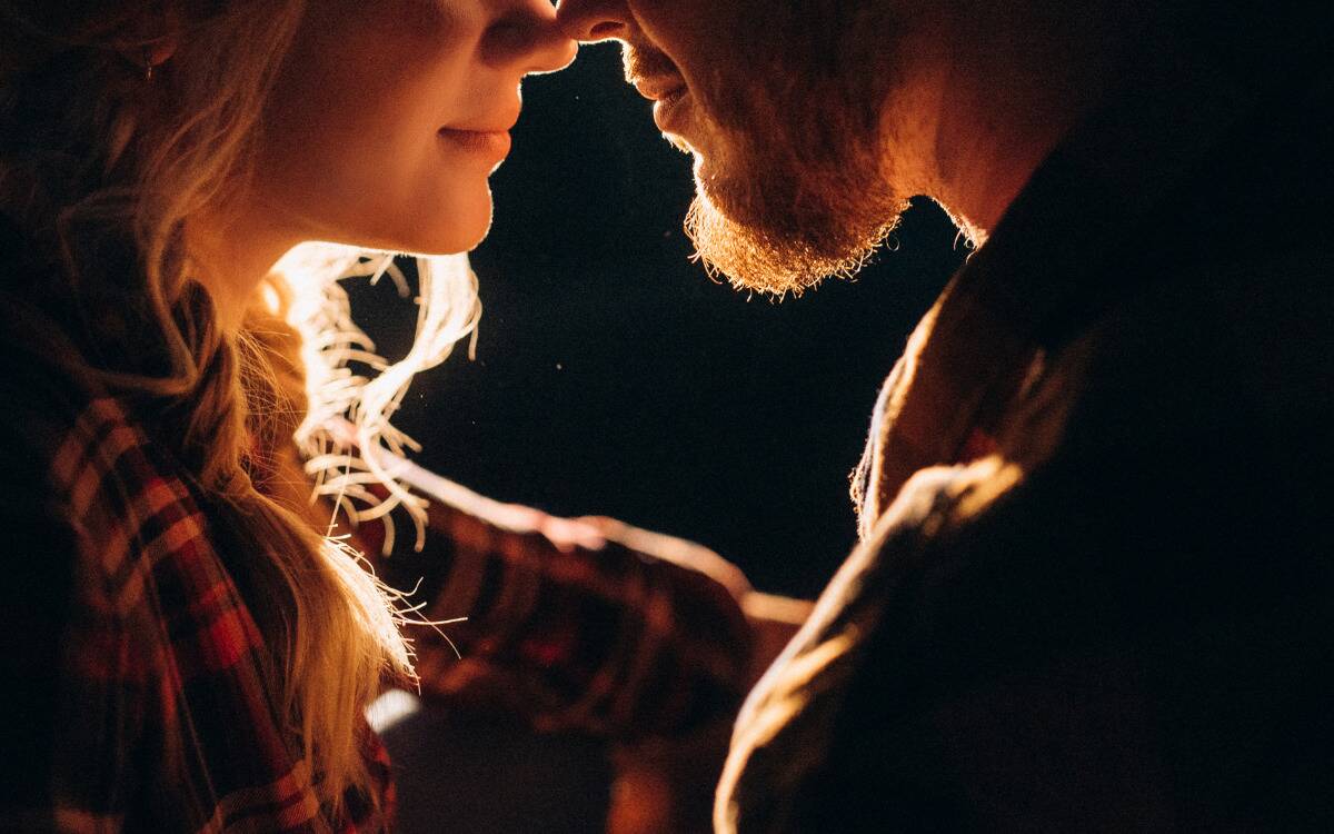 A closeup of a couple's faces, pressed close to one another, backlit in the dark.