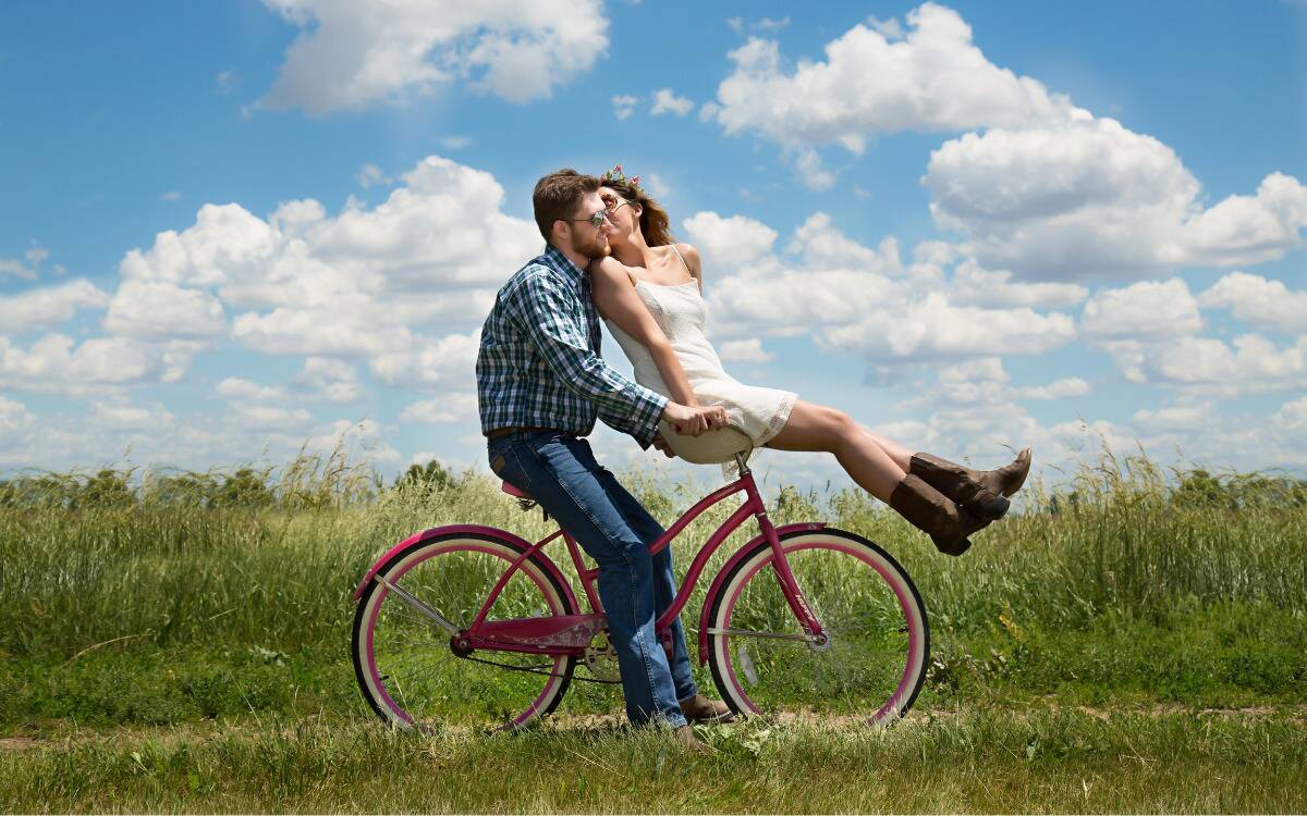 A man sitting on a bike on a dirt path next to a field. A woman is sitting on the handlebars, leaning back to kiss the man's cheek.