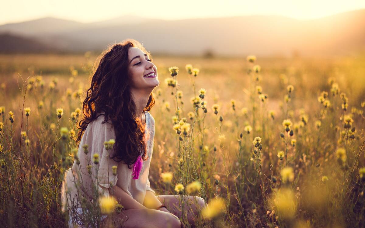 A woman smiling, eyes closed, sitting in a field of flowers.