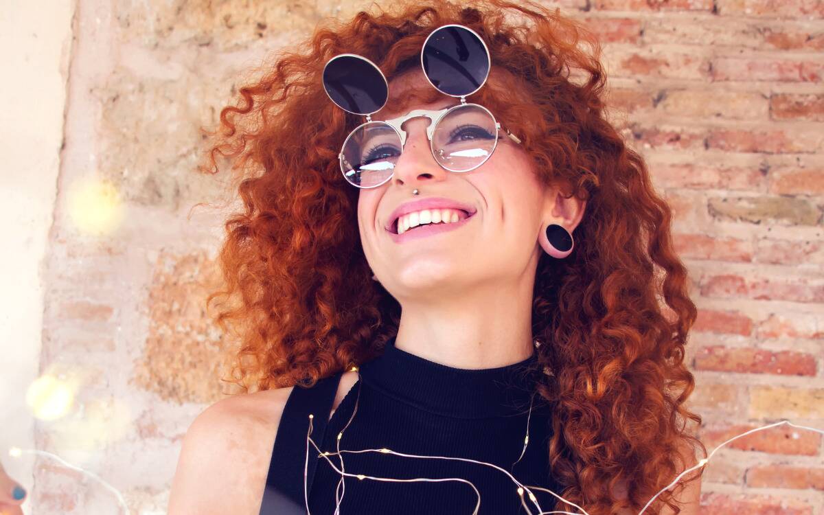 A woman with large, curly red hair and round sunglasses, smiling as she looks up.